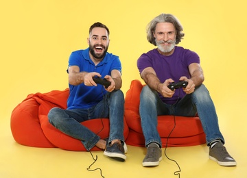 Mature man and guy playing video games with controllers on color background