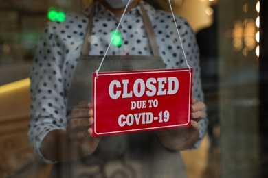 Woman in mask putting red sign with words Closed Due To Covid-19 hanging onto glass door, closeup