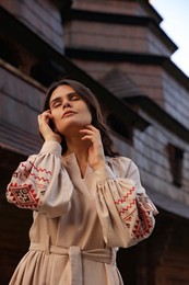 Photo of Beautiful woman wearing embroidered dress near old wooden church in village. Ukrainian national clothes