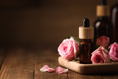 Bottles of essential rose oil and flowers on wooden table, space for text