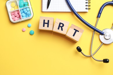 Wooden cubes with abbreviation HRT, pills, stethoscope and notebook on yellow background, flat lay. Hormone Replacement Therapy
