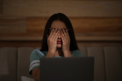 Young woman with nomophobia rubbing her eyes while using laptop at home late in evening. Insomnia concept