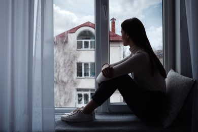 Melancholic young woman looking out of window indoors, space for text. Loneliness concept
