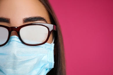 Young woman with foggy glasses caused by wearing disposable mask on pink background, space for text. Protective measure during coronavirus pandemic