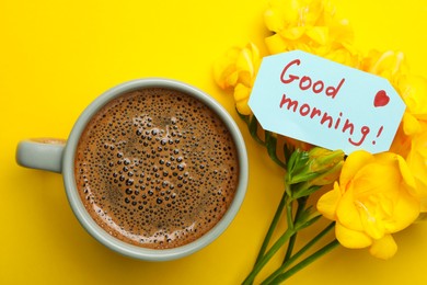 Cup of aromatic coffee, beautiful freesias and Good Morning note on yellow background, flat lay