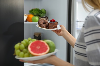 Photo of Choice concept. Woman holding plates with fruits and sweets near refrigerator in kitchen, closeup
