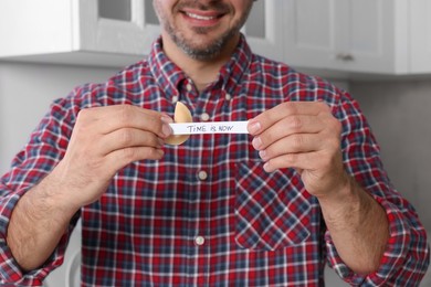 Photo of Happy man holding tasty fortune cookie with prediction Time is Now indoors, closeup