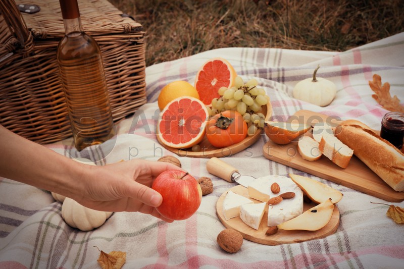 Woman taking apple from blanket with snacks and wine outdoors, closeup. Autumn picnic
