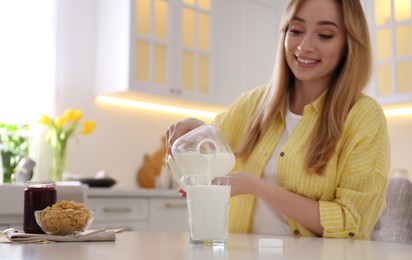 Young woman pouring milk from gallon bottle into glass at white table in kitchen