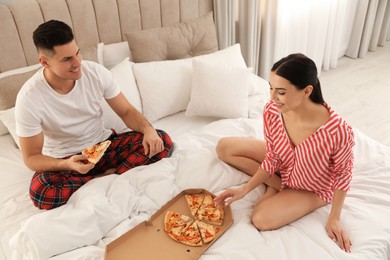 Happy couple in pyjamas eating pizza on bed at home