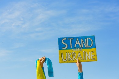Photo of Woman holding poster Stand with Ukraine and national flag against blue sky, closeup