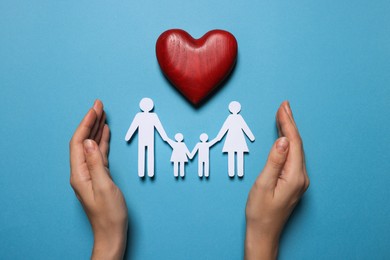 Photo of Woman protecting paper family figures and red wooden heart on light blue background, top view. Insurance concept