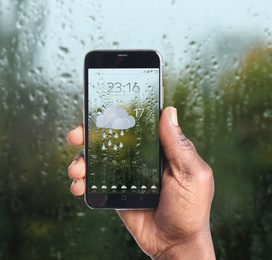 Image of African-American man using weather forecast app on smartphone near window indoors, closeup