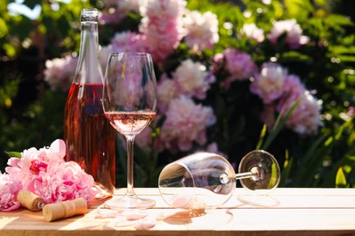 Bottle and glass of rose wine near beautiful peonies on wooden table in garden. Space for text