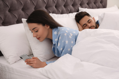 Young woman using smartphone while her boyfriend sleeping in bed at home