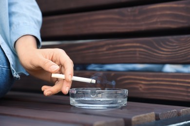 Woman holding cigarette over glass ashtray on bench outdoors, closeup