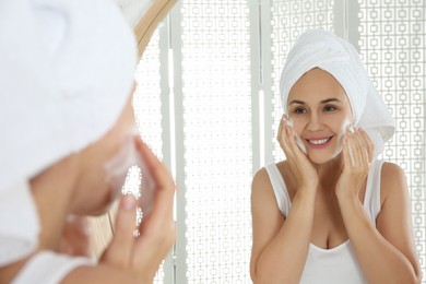 Happy mature woman applying cleansing foam onto face near mirror in bathroom