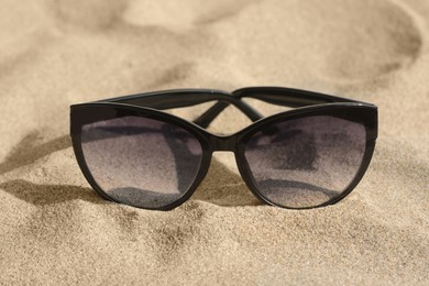 Photo of Stylish sunglasses with black frame on sandy beach, closeup