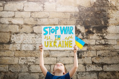 Boy holding poster Stop War In Ukraine and national flag against brick wall outdoors