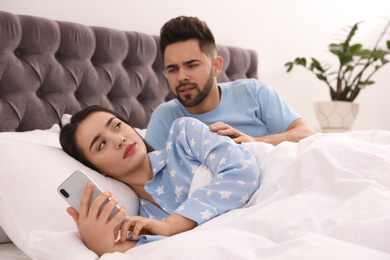 Young woman ignoring her distrustful boyfriend while using smartphone in bed at home