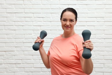 Happy overweight woman doing exercise with dumbbells near white brick wall, space for text