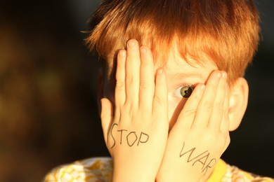 Little boy hiding face and words Stop War written on his hands outdoors, closeup