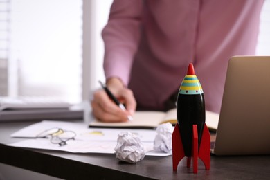 Photo of Woman working at messy table, focus on toy rocket. Startup concept