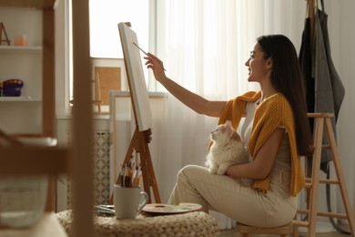 Beautiful young woman drawing on easel with cat at home
