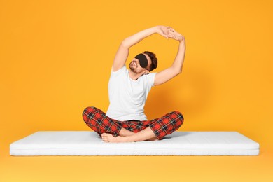 Man in sleeping mask sitting on soft mattress and stretching against orange background