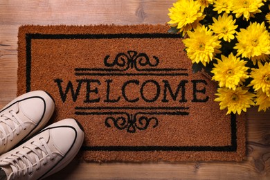 Photo of Doormat with word Welcome, stylish sneakers and beautiful flowers on wooden floor, flat lay