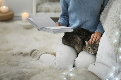 Young woman with cute cat reading book at home, closeup. Cozy winter