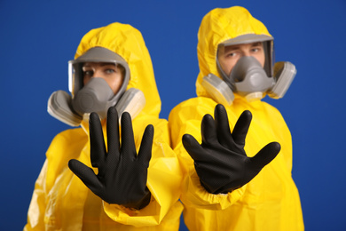 Man and woman in chemical protective suits making stop gesture against blue background, focus on hands. Virus research