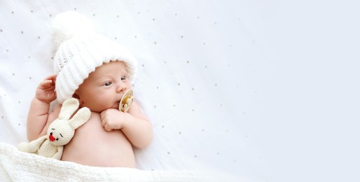 Cute newborn baby in white knitted hat with toy lying on bed, top view with space for text. Banner design