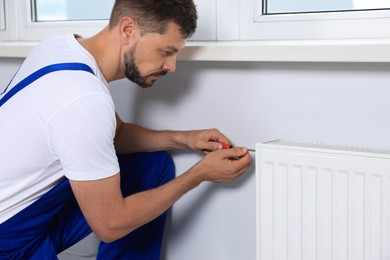 Professional plumber using screwdriver while preparing heating radiator for winter season indoors