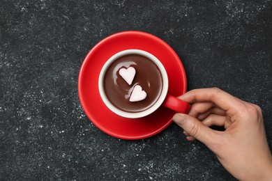 Photo of Woman holding cup of hot chocolate with heart shaped marshmallows at grey table, top view