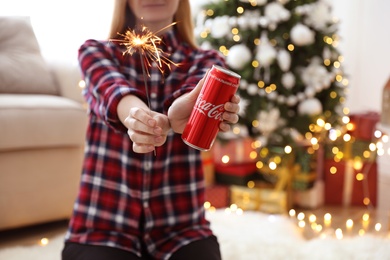 MYKOLAIV, UKRAINE - January 01, 2021: Woman with can of Coca-Cola and sparkler against blurred Christmas tree at home, closeup