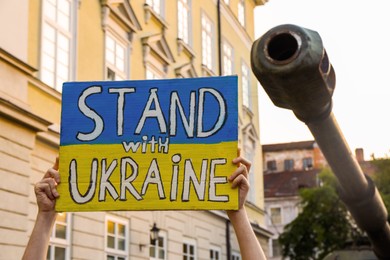 Woman holding poster in colors of national flag and words Stand With Ukraine near broken military tank outdoors, closeup