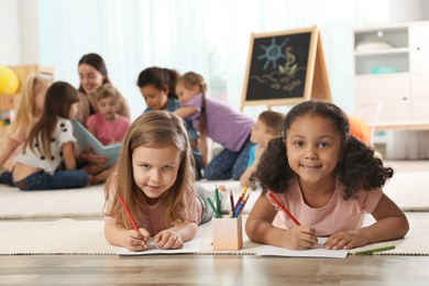 Cute girls drawing on floor while kindergarten teacher reading book to other children indoors