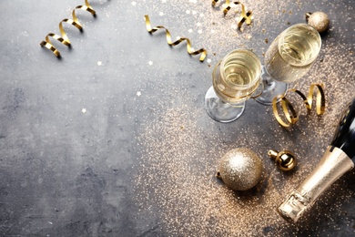 Glasses and bottle of champagne, serpentine streamers with Christmas balls on grey table, above view. Space for text
