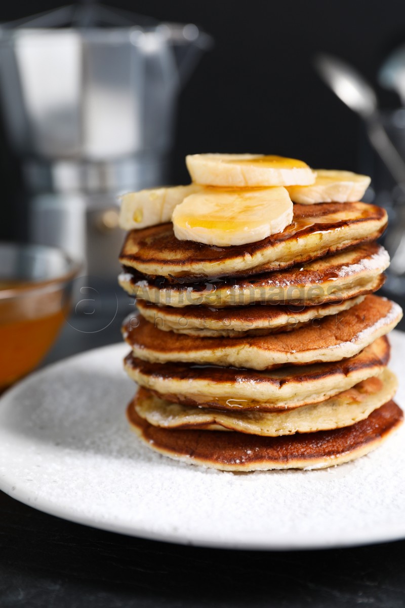 Photo of Plate of banana pancakes with honey and powdered sugar on black table, closeup