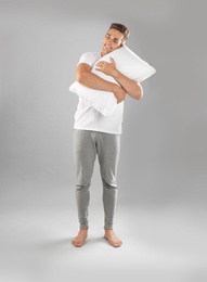 Young man in pajamas embracing pillow on gray background