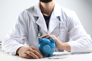 Doctor with piggybank and money at white table indoors, closeup. Medical insurance concept