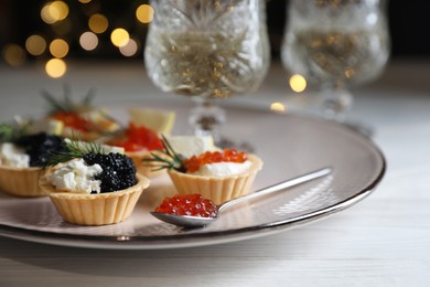 Photo of Delicious tartlets with red and black caviar served on white wooden table against blurred festive lights, closeup