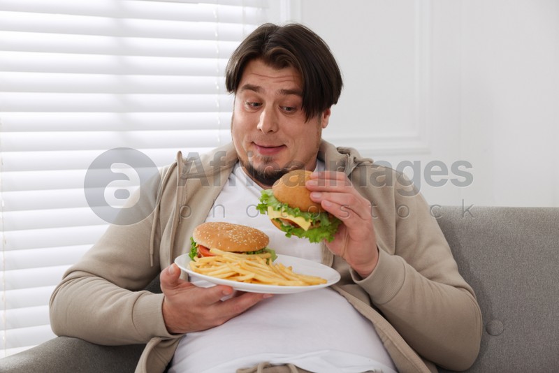 Overweight man with plate of burgers and French fries on sofa at home