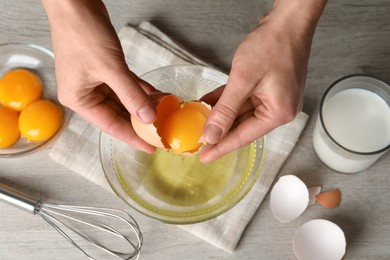 Woman separating egg yolk from white over glass bowl at light wooden table, top view
