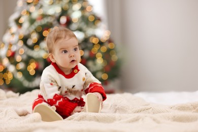 Cute little baby in Christmas sweater on knitted blanket against blurred festive lights, space for text. Winter holiday