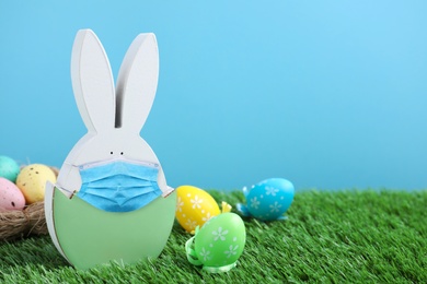 COVID-19 pandemic. Easter bunny figure in protective mask and dyed eggs on green grass against light blue background, space for text