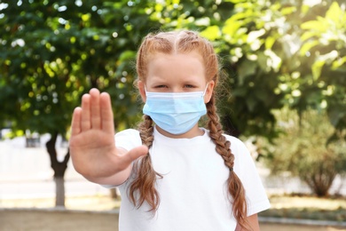 Little girl in protective mask showing stop gesture outdoors. Prevent spreading of coronavirus