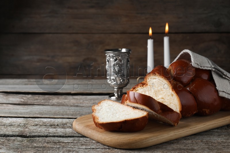 Cut homemade braided bread, goblet and candles on wooden table, space for text. Traditional Shabbat challah