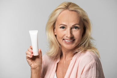 Portrait of beautiful mature woman with perfect skin holding tube of cream on grey background